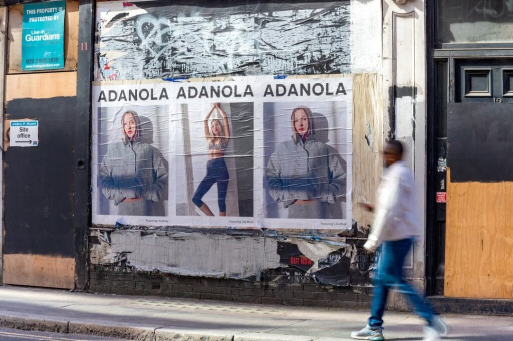 Wall of fashion posters reading 'ADANOLA' repeated three times, featuring a woman in a gray hoodie in each panel; a blurred passerby walks by on a city sidewalk behind damaged storefronts.
