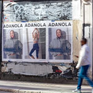 Wall of fashion posters reading 'ADANOLA' repeated three times, featuring a woman in a gray hoodie in each panel; a blurred passerby walks by on a city sidewalk behind damaged storefronts.